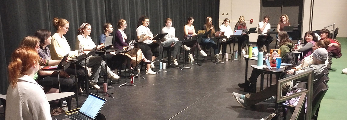 Cast of (&Medea) sits on stage in a semicircle, in front of a large black curtain.